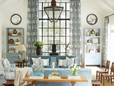 Living room with vaulted ceiling, white paneled walls, and a large black-paned window. Features a light blue sofa with patterned pillows, two blue bookshelves, and a patterned armchair.