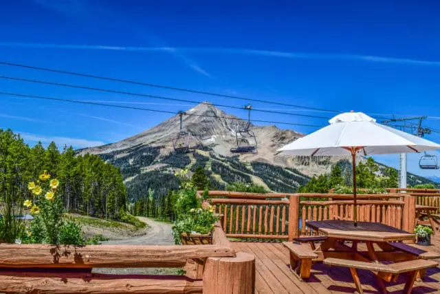 Wooden deck with picnic tables and umbrella overlooking a mountain with ski lifts and trees under a bright blue sky