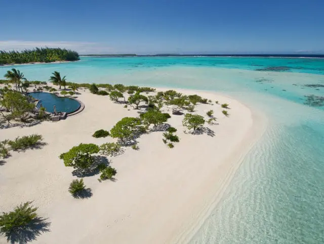 Aerial view of a tropical island with a curved infinity pool, white sand beach, lush green trees, and clear turquoise water under a bright blue sky.