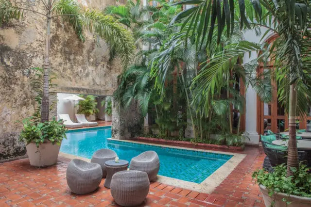Courtyard with a turquoise tiled swimming pool, surrounded by lush palm trees, terracotta brick paving, and wicker seating.