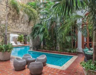 Courtyard with a bright blue swimming pool, surrounded by lush tropical plants and palm trees. A seating area with four grey woven chairs and a small table sits on a terracotta tile patio.
