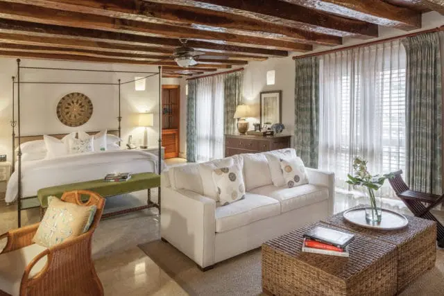 Bedroom with a four-poster bed, white sofa, wicker coffee table, and exposed wooden ceiling beams. Dominant colors are white, green, and natural wood tones.