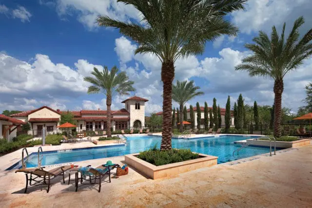 Resort-style swimming pool with lounge chairs and umbrellas, surrounded by palm trees and Mediterranean-style architecture under a partly cloudy sky.