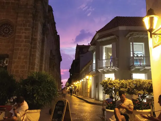 Cobblestone street lined with colonial-style buildings and gas lamps under a vibrant purple and pink sunset sky. People sit at outdoor cafes.
