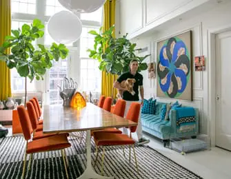 Man holding a small dog in a dining room with orange chairs, a teal sofa, large plants, and abstract art on the wall.