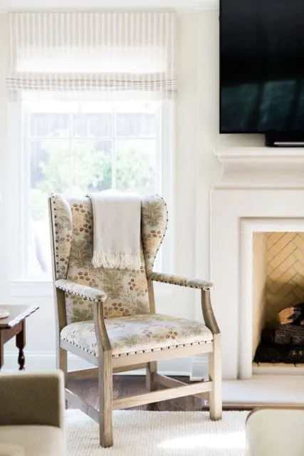 Wingback armchair with botanical print upholstery and nailhead trim, draped with a fringed throw blanket, next to a window with striped Roman shades and a fireplace with herringbone brick surround.