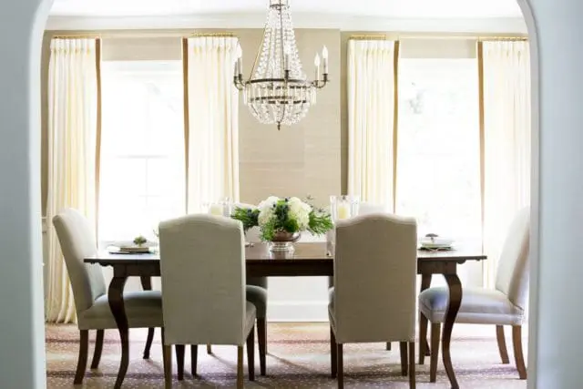 Dining room with a dark wood table set with place settings and a floral centerpiece, surrounded by upholstered chairs. Cream curtains frame windows, and a crystal chandelier hangs above.