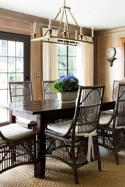 Dining room with dark wood table, rattan chairs with white cushions, and a rectangular brass chandelier. A blue hydrangea centerpiece sits on the table.