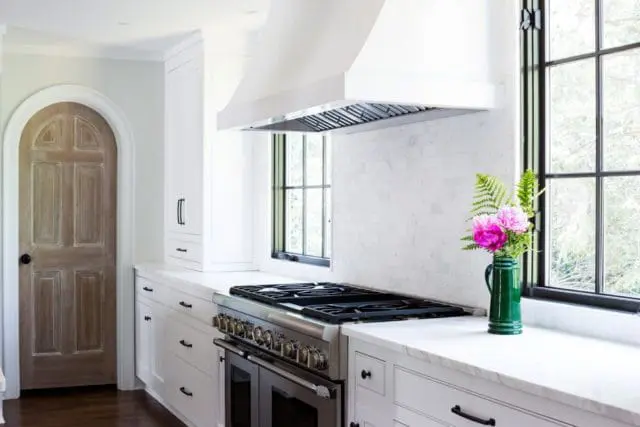 White kitchen with arched wooden door, marble countertops, stainless steel range, and white subway tile backsplash. A green pitcher holds pink peonies and ferns.