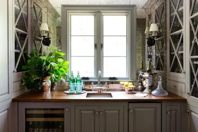 Bar area with gray cabinetry, wood countertop, and mirrored walls. Features a sink, wine fridge, bottles of water, lemons, and champagne buckets.