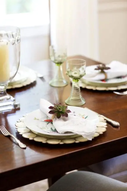 Dining table set with floral plates, white napkins adorned with succulents, green stemmed glassware, and a tall glass candle holder.
