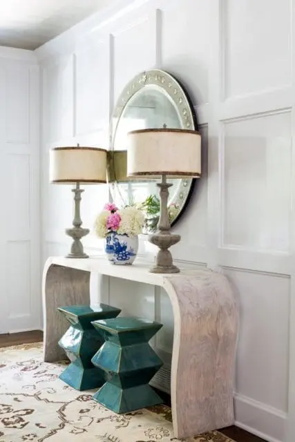 Entryway with paneled walls, a light wood console table topped with two lamps, a blue and white vase of flowers, and a round mirror. Two teal garden stools sit below the table.