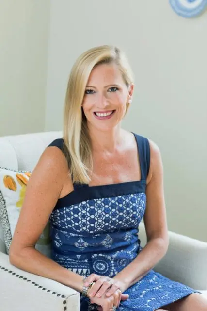Blonde woman with blue eyes wearing a blue lace dress and pearl earrings, sitting in a cream armchair with a patterned pillow.