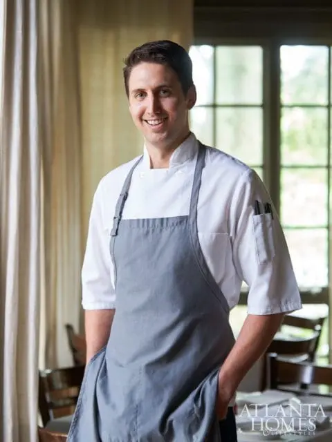 Chef in white chef coat and grey apron with hands in pockets, smiling at the camera. Atlanta Homes & Lifestyles logo in the bottom right corner.