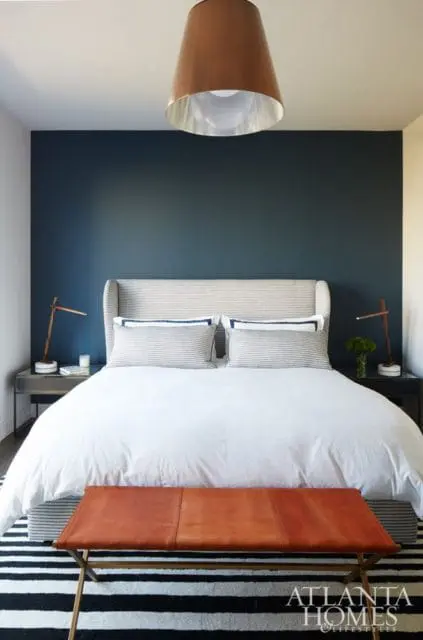 Bedroom with a dark blue accent wall, king-size bed with white linens and striped pillows, a copper pendant light, and a leather bench on a striped rug.