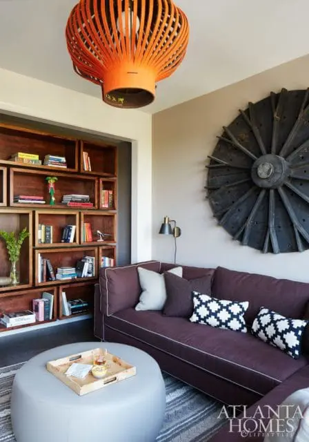 Living room with a plum sofa, patterned throw pillows, a light gray ottoman with a tray, and a large wooden wall art.