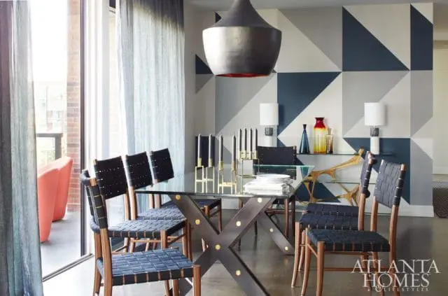 Dining room with glass-top table, dark metal X-base, and black woven chairs. Geometric blue and gray wallpaper accents the wall.