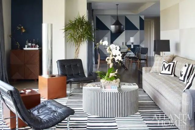 Living room with black leather chairs, a striped ottoman coffee table, and a patterned sofa. A geometric mural is visible in the background.