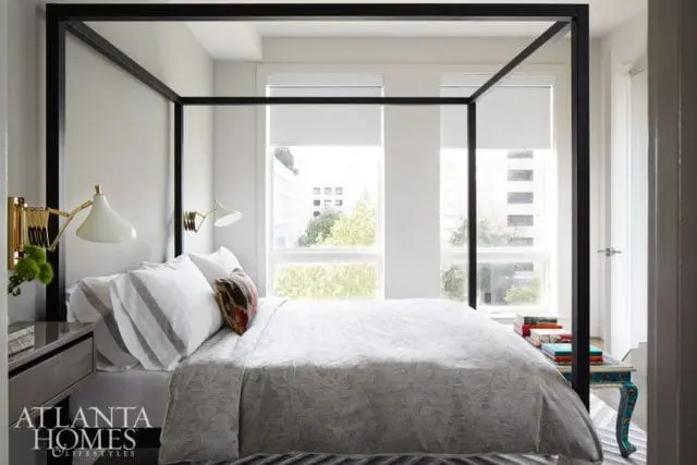Bedroom with a black canopy bed, white bedding, and gold wall sconces. Large windows overlook a city view.