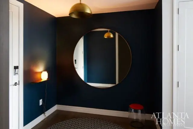 Hallway with dark blue walls, a large round mirror, a brass pendant light, and a small stool with a red top.
