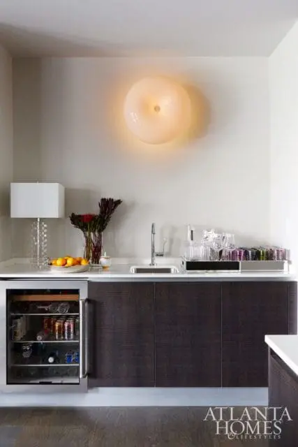 Modern wet bar with dark wood cabinetry, stainless steel wine fridge, white countertop, sink, and a circular wall sconce.