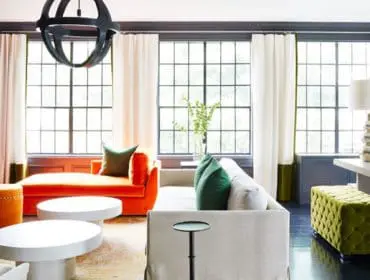 Living room with dark walls, large grid windows, orange chaise lounge, white sofa, green tufted ottoman, and white coffee tables.