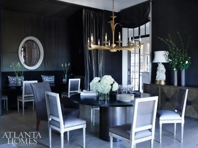 Dark dining room with a round black table set for dinner, upholstered chairs with white frames, a gold chandelier, and a mirrored oval wall hanging.