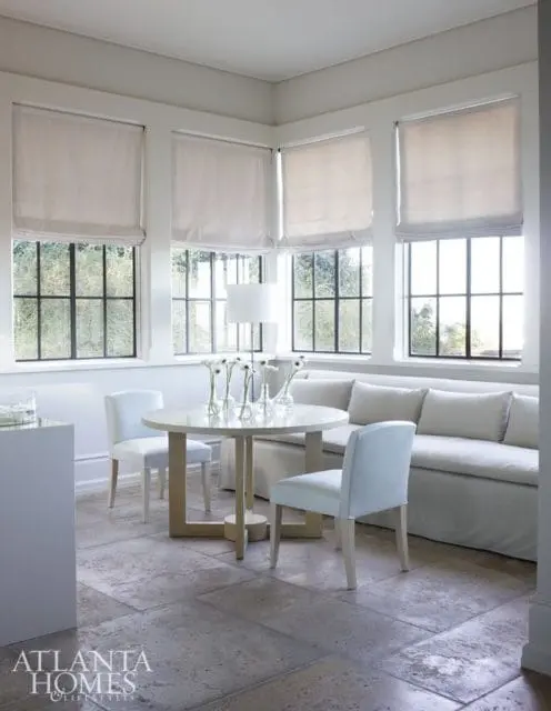 Light-filled kitchen nook with a round dining table, two chairs, and a built-in banquette seating. Large windows with Roman shades overlook greenery.