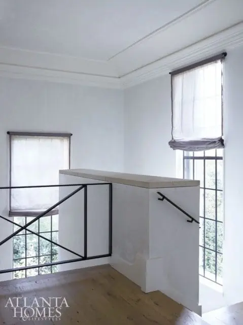 Interior view of a staircase landing with white walls, a stone-topped railing, black metal balusters, and large windows with sheer Roman shades.