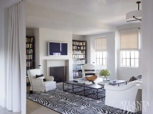 Living room with white armchairs, a zebra print rug, dark coffee tables, and bookshelves flanking a fireplace with a mounted television.