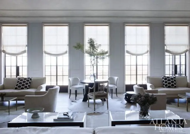 Living room with neutral-toned sofas and armchairs, a round dining table with chairs, and large windows with Roman shades.