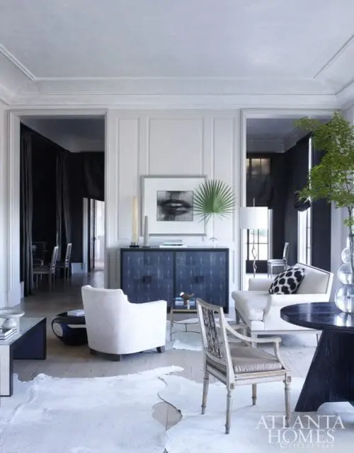Living room with white upholstered chairs, a dark blue textured cabinet, and a black round table. A large white hide rug covers the light wood floor.