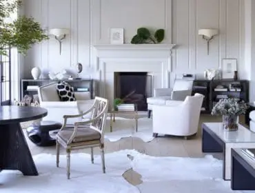 Living room with white paneled walls, fireplace, white sofas, armchairs, a dark wood dining table with a glass vase of greenery, and cowhide rugs.