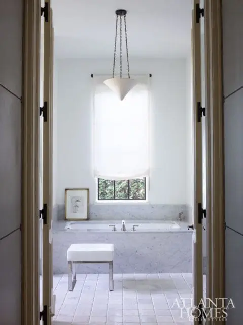 White bathroom with marble bathtub surround, white tiled floor, a white upholstered bench, and a pendant light fixture.