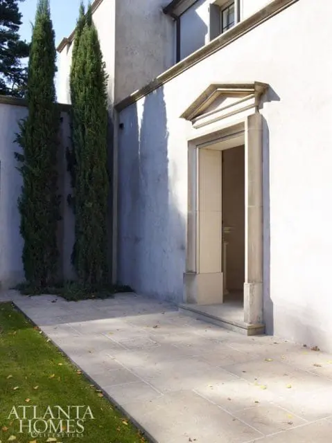 Exterior of a building with a light stucco finish, featuring a stone entryway with a pediment detail, flanked by tall cypress trees and a stone patio.