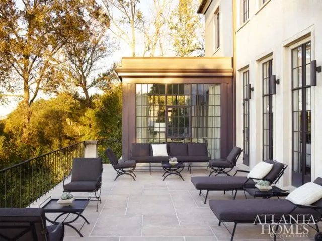 Large stone patio with dark upholstered outdoor furniture, including sofas, chairs, and chaise lounges, overlooking a wooded hillside. A large windowed sunroom is visible on the house.