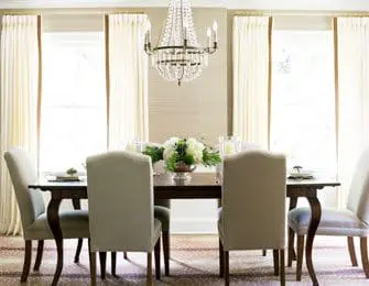 Dining room with dark wood table, upholstered chairs, crystal chandelier, and tall cream curtains framing windows.