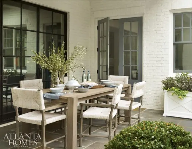 Covered patio with a light wood dining table and chairs set for a meal, white brick walls, and large dark-framed windows.