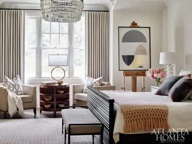 Bedroom with a large window, two armchairs, a round side table, a bed with a fringed throw, and abstract art on an easel.
