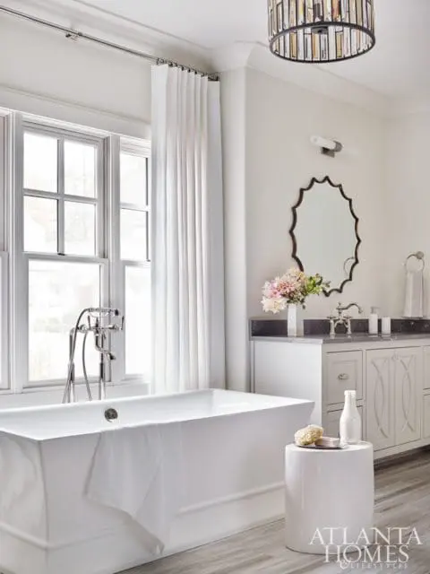 White freestanding bathtub with chrome fixtures next to a window with white curtains, and a white vanity with a dark countertop and decorative mirror above.