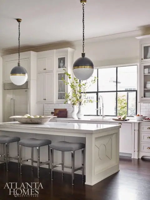 White kitchen with marble island, gray upholstered stools, and globe pendant lights. Stainless steel refrigerator and white cabinetry are visible.