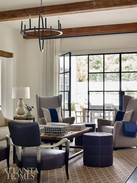 Living room with wingback chairs, leather armchairs, a rustic wood coffee table, and a large black chandelier. Exposed wooden beams accent the ceiling.