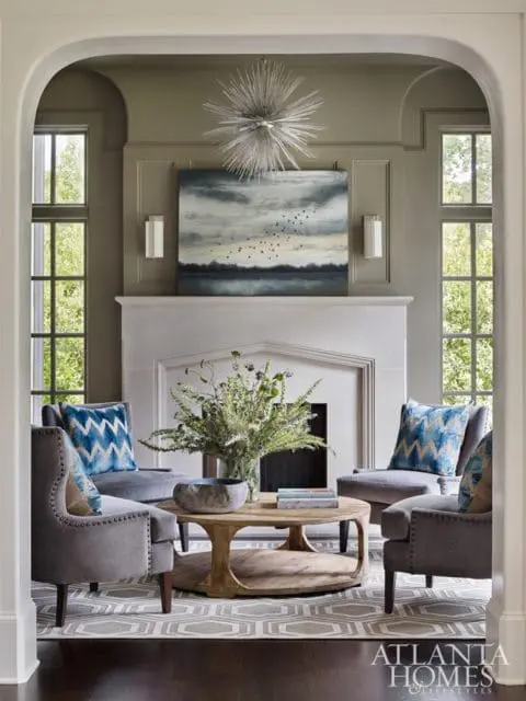 Living room with gray upholstered chairs, a light stone fireplace, a large abstract painting of birds over water, and a wooden coffee table with a floral arrangement.
