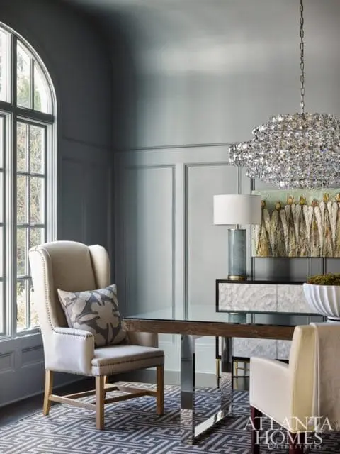 Gray paneled dining room with a tall wingback chair, glass-top dining table with chrome legs, abstract art, and a crystal chandelier.