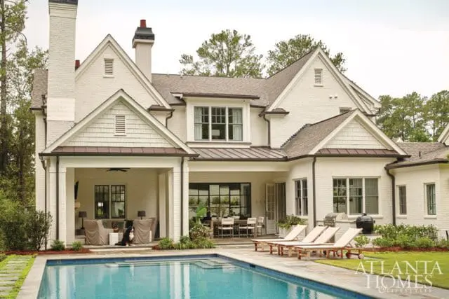 White brick house with dark trim, multiple gables, and a covered porch overlooking a swimming pool and patio with lounge chairs.