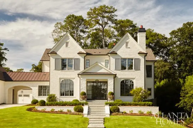 White brick house with gray shutters, arched windows, and a copper roof over the garage. Landscaped lawn and trees surround the property.