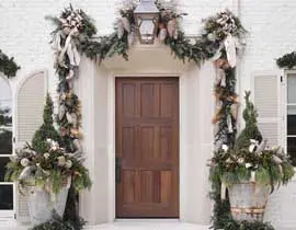 Front door of a white brick house decorated for the holidays with a large wooden door, garland, urns filled with greenery, and shutters.