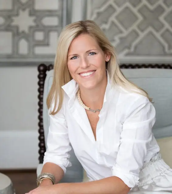 Ansley Ghegan smiling, wearing a white button-down shirt and a statement necklace, seated in a chair with a decorative wall behind her.