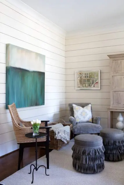 Living room corner with shiplap walls, featuring a large abstract painting, a wicker chair, a grey armchair with a patterned pillow, and two grey ottomans with fringe detail.