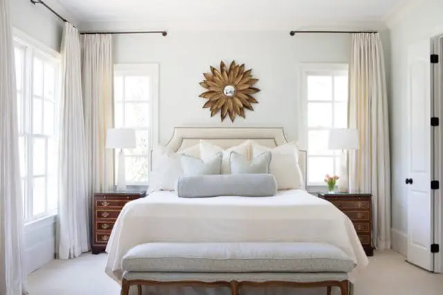 Bedroom with a king-size bed featuring a tufted headboard, white bedding, and light blue accent pillows. A gold sunburst mirror hangs above the bed.